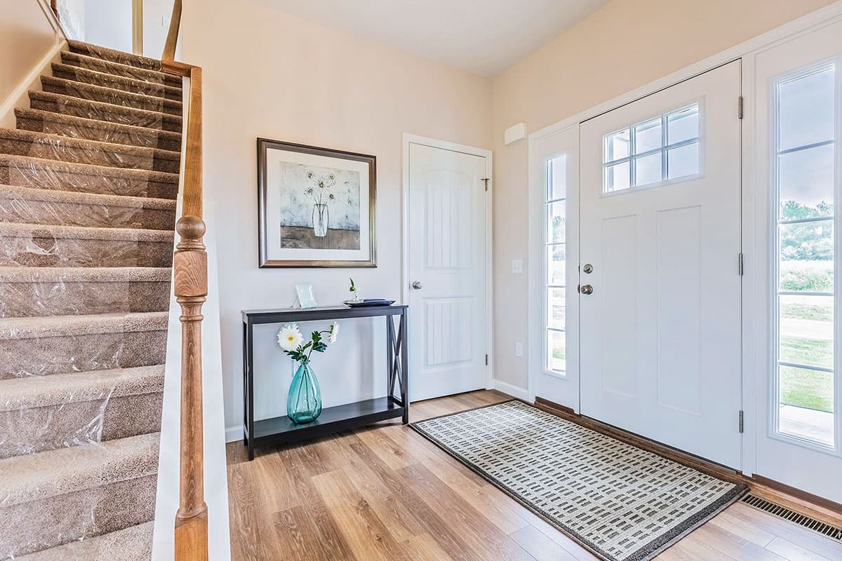 Interior view of a house entryway with carpeted stairs, a console table with vase, and a white front door with sidelights.