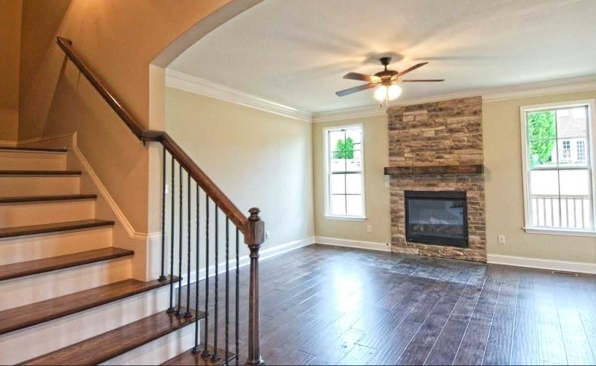 Interior view of a living room featuring a stone fireplace, dark wood floors, a staircase with wrought iron spindles, and a ceiling fan.