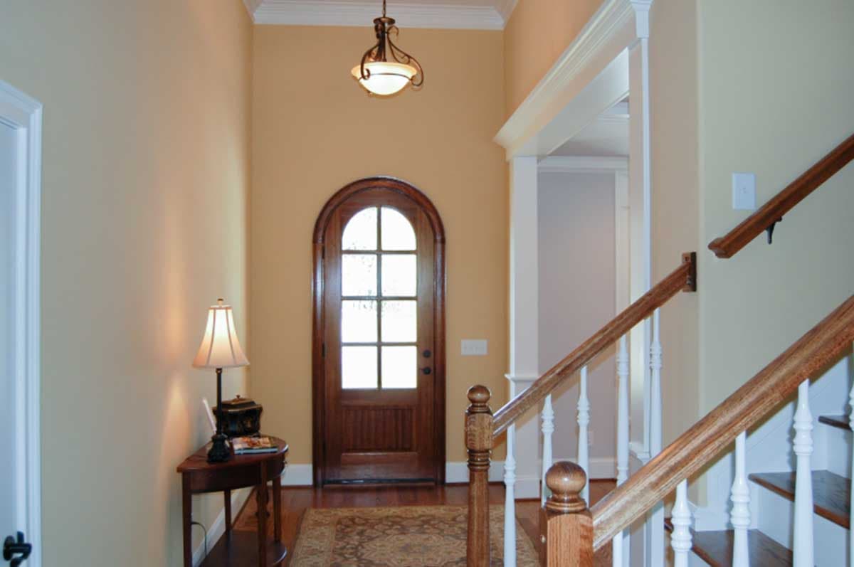 Foyer with arched wooden door with glass panes, staircase with wooden handrail, and decorative pendant light.