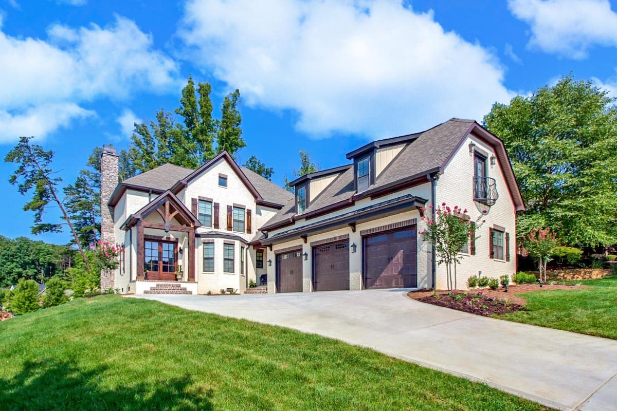 House plan exterior: Two-story white brick home with a prominent stone chimney, wooden gabled entryway, dormers, and a three-car garage.