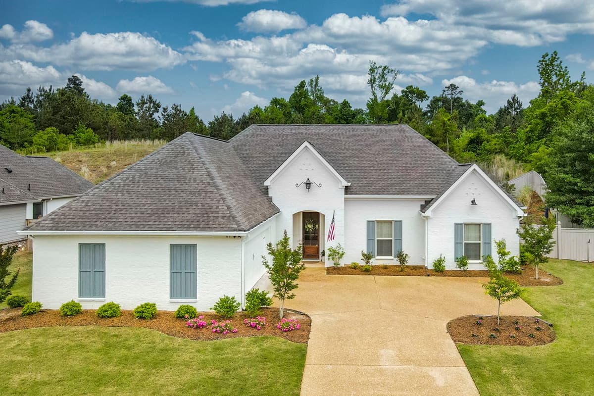House plan exterior. One-story house in white brick with gabled roof, shutters, and an arched entry. Features a driveway and manicured lawn.