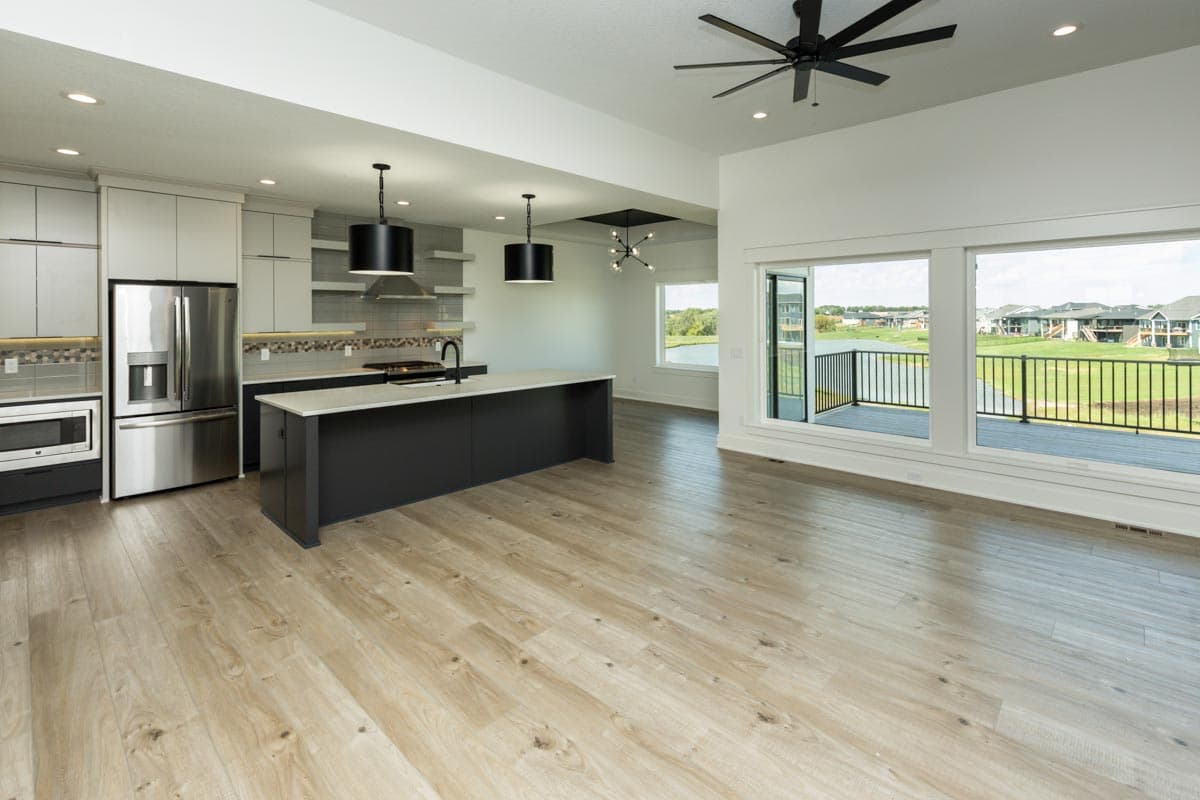 Modern kitchen with island and stainless steel appliances adjacent to great room with vaulted ceiling and large windows overlooking a lake.