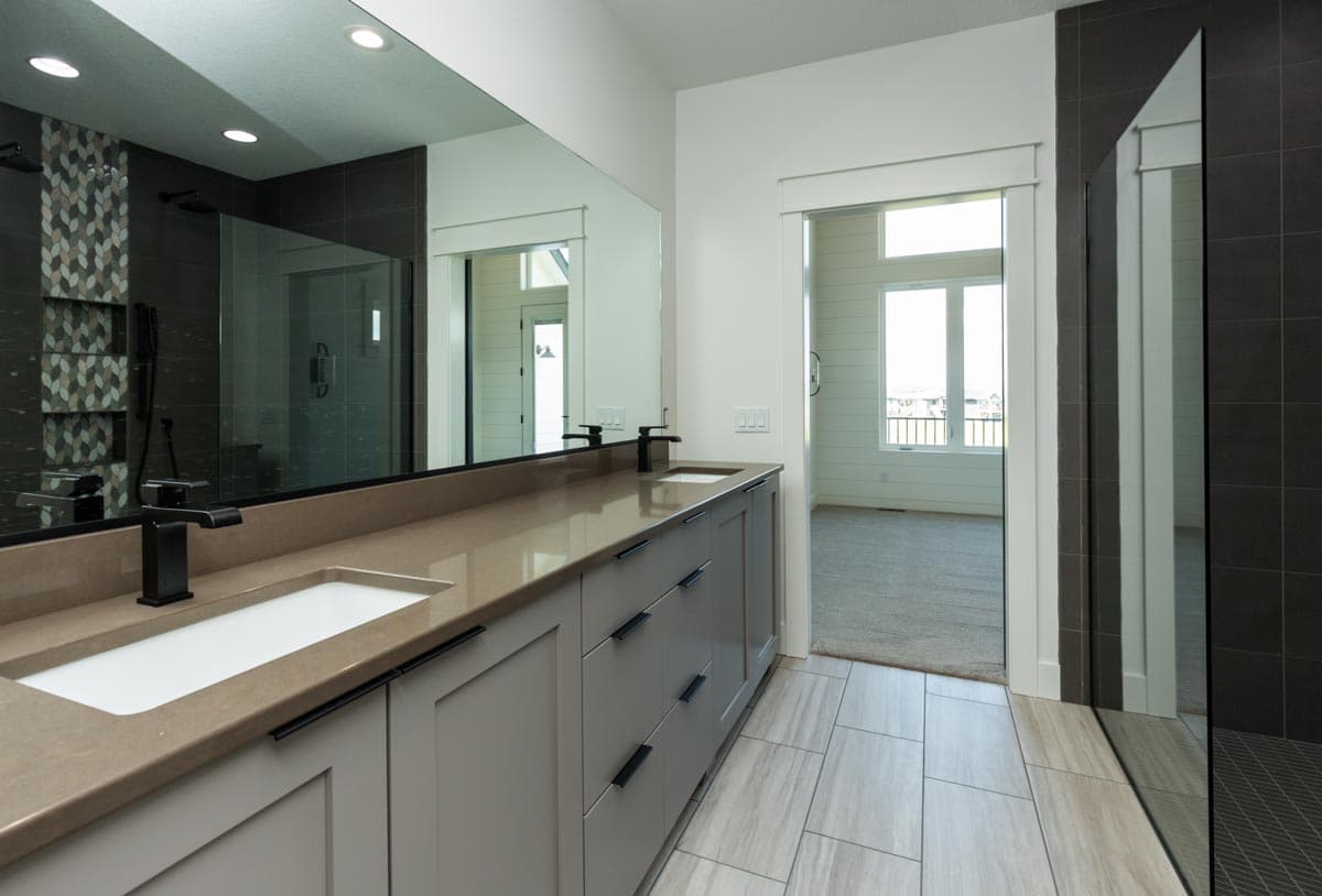 Bathroom with double vanity, gray cabinetry, tan countertop, separate shower with tile detail, and view into a bedroom.