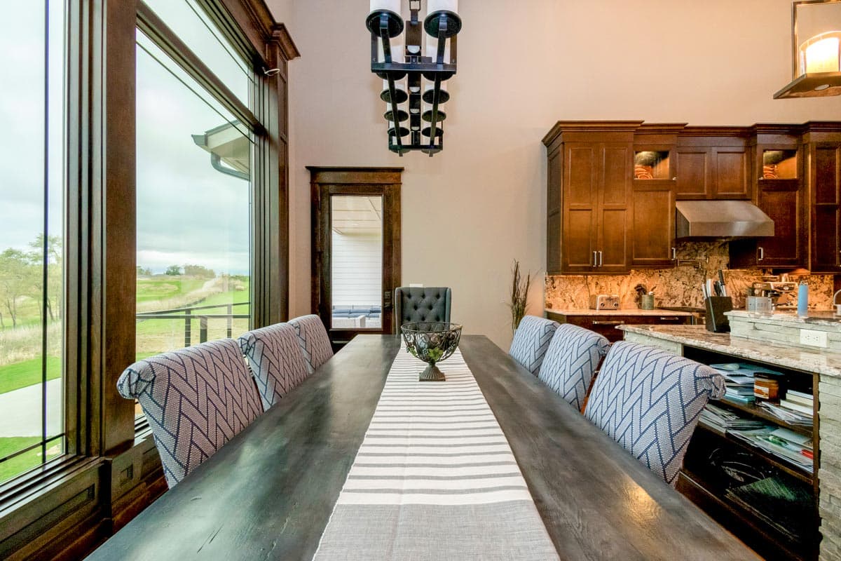 Dining area with long table, patterned chairs, wall of windows, and adjacent kitchen with dark wood cabinets and granite countertops.
