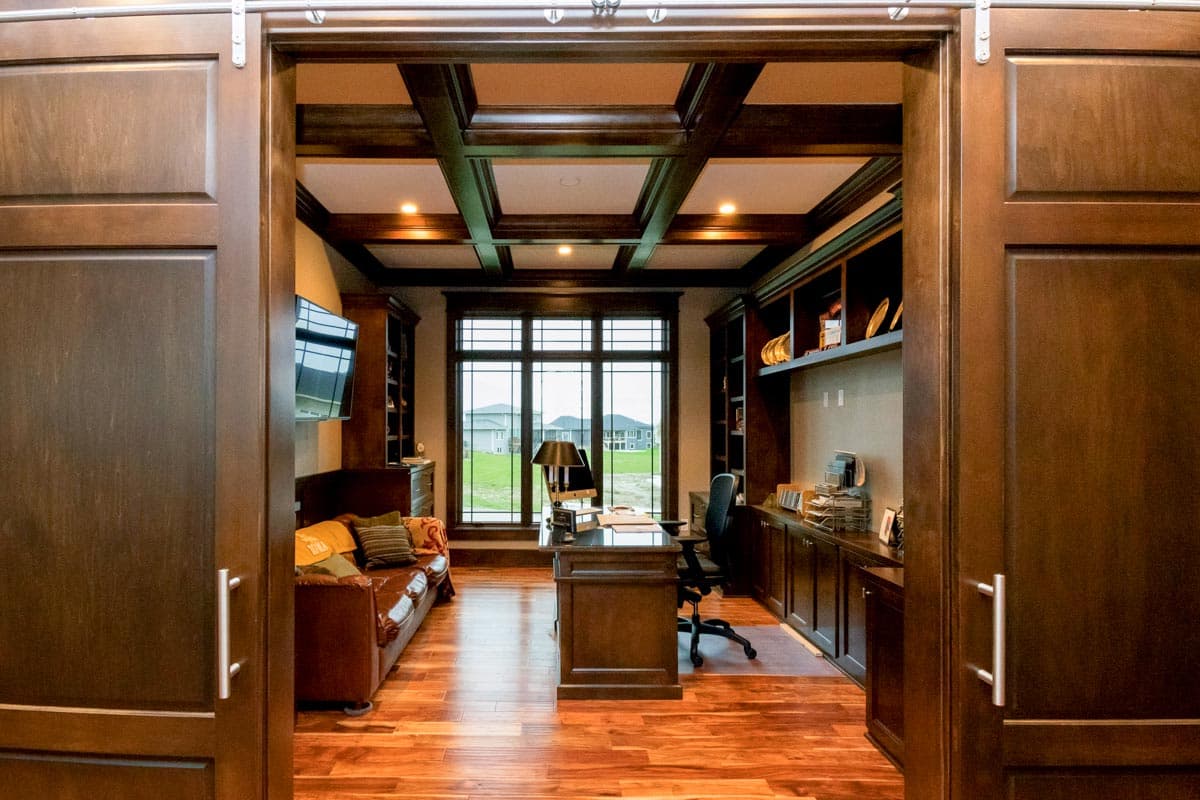 Home office with dark wood built-in bookshelves, coffered ceiling, leather sofa, desk, and large window.