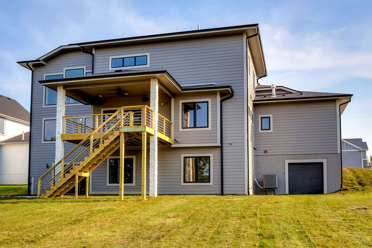 House plan exterior: Modern style, two stories, gray siding, covered deck with stairs, garage, and multiple window configurations.