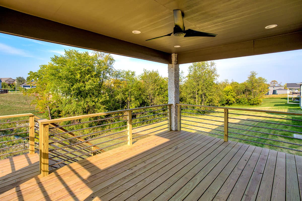 Covered deck with wood flooring, ceiling fan, recessed lighting, and horizontal cable railings overlooking a grassy yard.