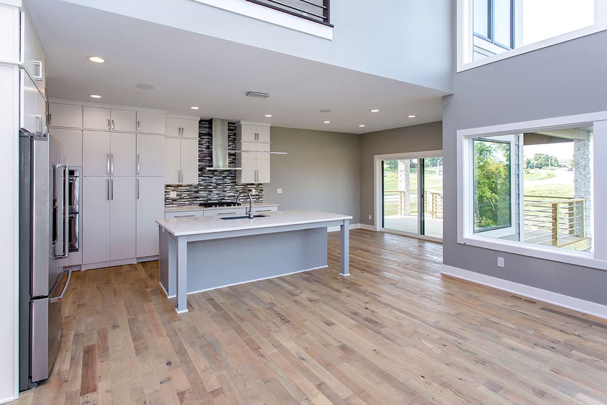 Open concept living area with a modern kitchen featuring an island, and large windows overlooking an outdoor deck. Two-story ceiling.
