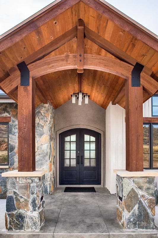 Exterior view of house plan with wood beam portico, stone base pillars, arched double doors, and multi-pane windows.