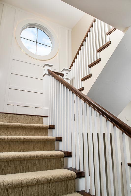 Carpeted staircase with white balusters, dark wood handrail, and a circular window above paneled wainscoting.