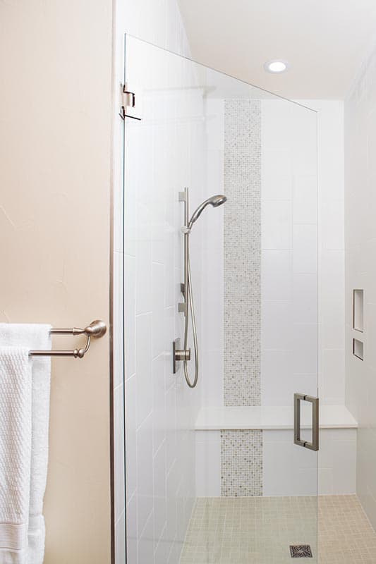 Shower stall with glass door, white tile walls, mosaic accent, built-in bench, handheld shower head, and towel bar.