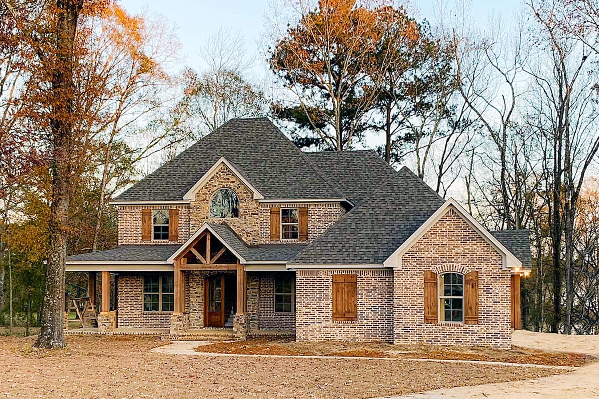 House plan exterior. Two-story brick home with a covered porch, gabled roof, and decorative wooden shutters.