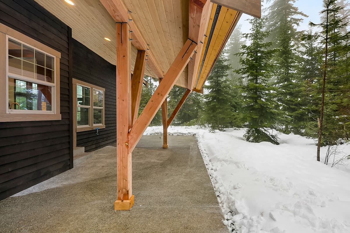House plan exterior showing dark horizontal siding, multiple windows, and exposed wood structural supports for a covered patio in a snowy environment.