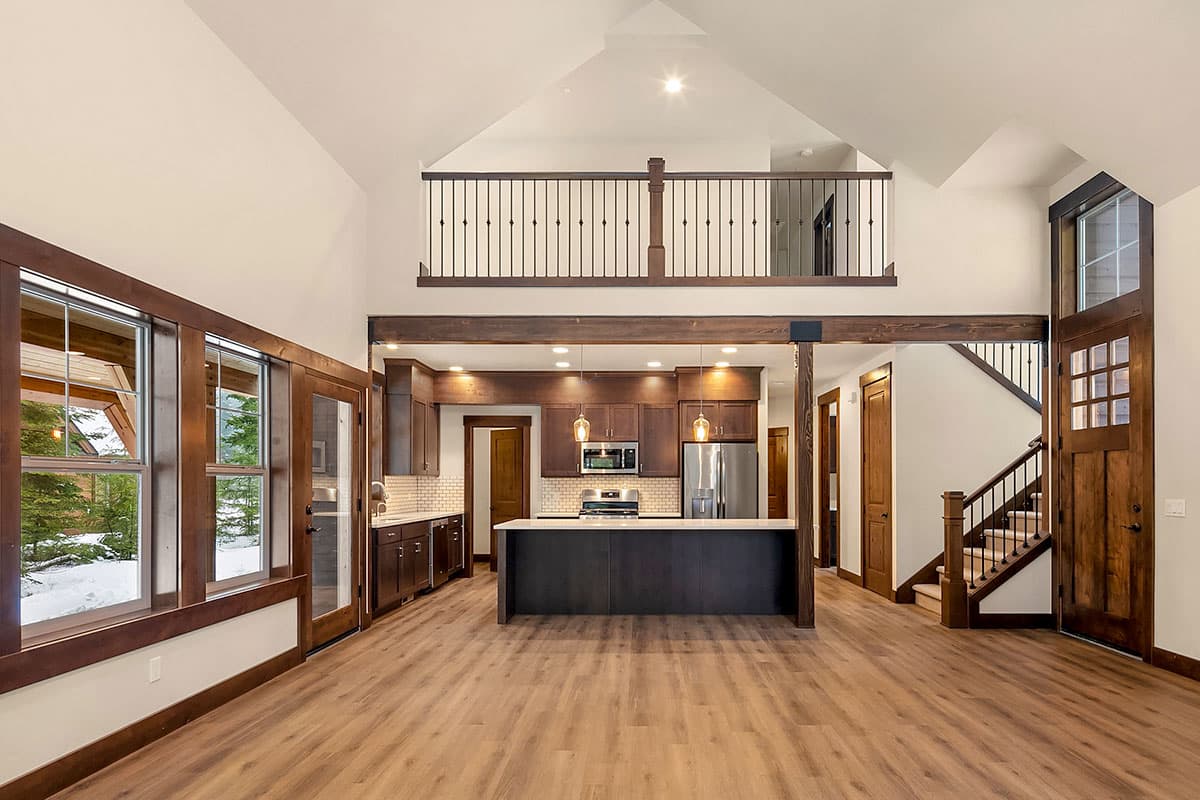 Interior view of a great room with vaulted ceiling, exposed beams, large windows, a kitchen island, and a loft overlook.