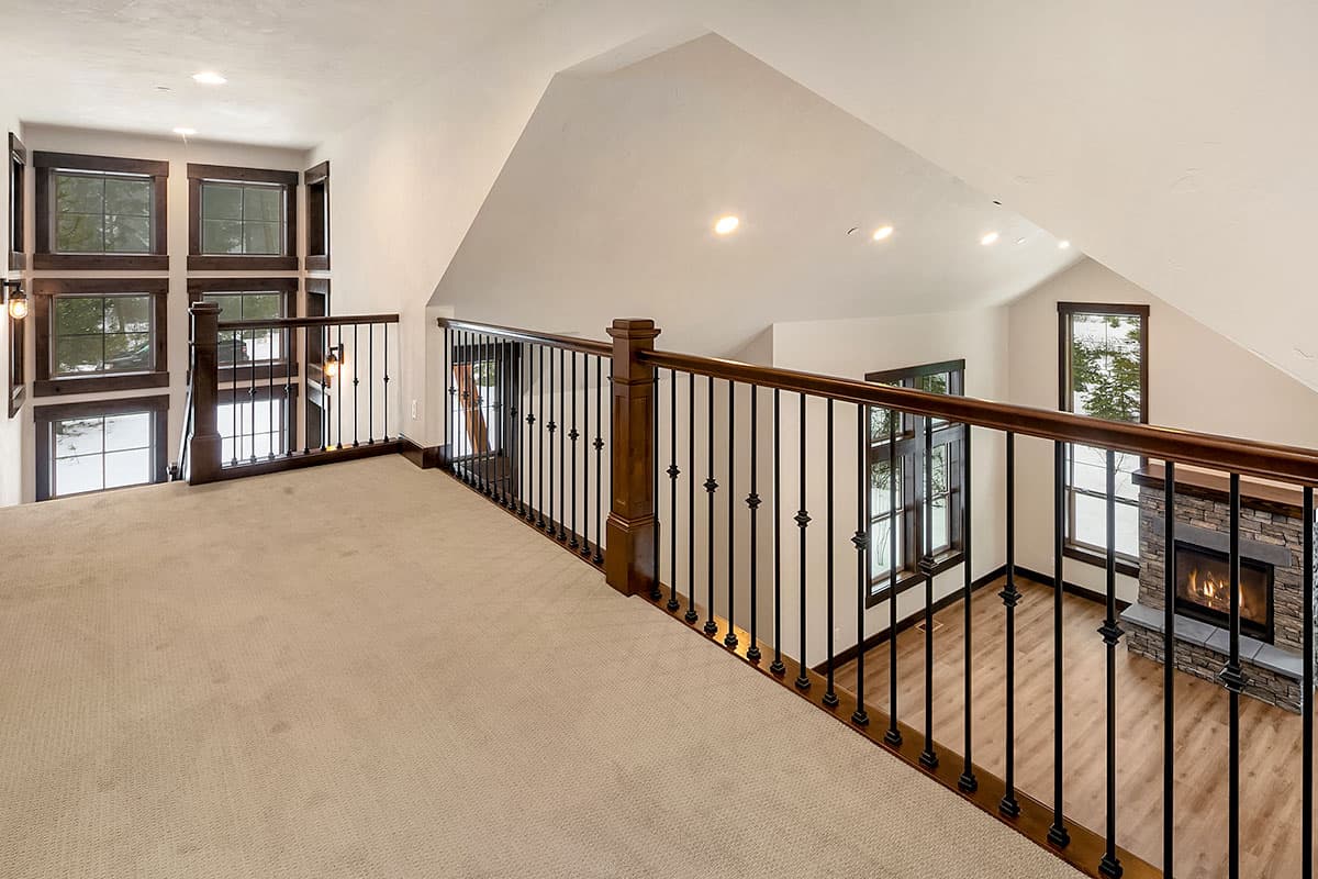 Interior view of a loft overlooking a living area with stone fireplace, vaulted ceiling, and large windows.