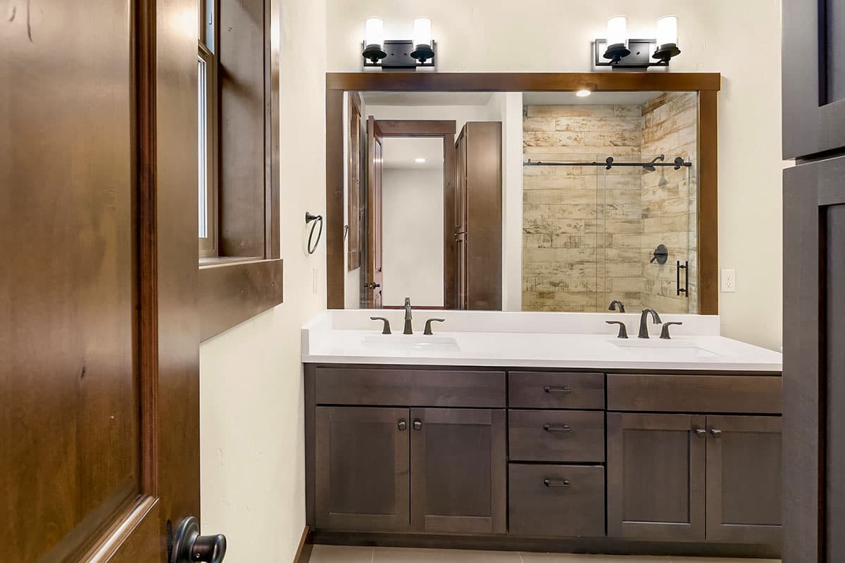 Bathroom vanity with double sinks and dark wood cabinets, reflected in mirror showing wood-plank shower.
