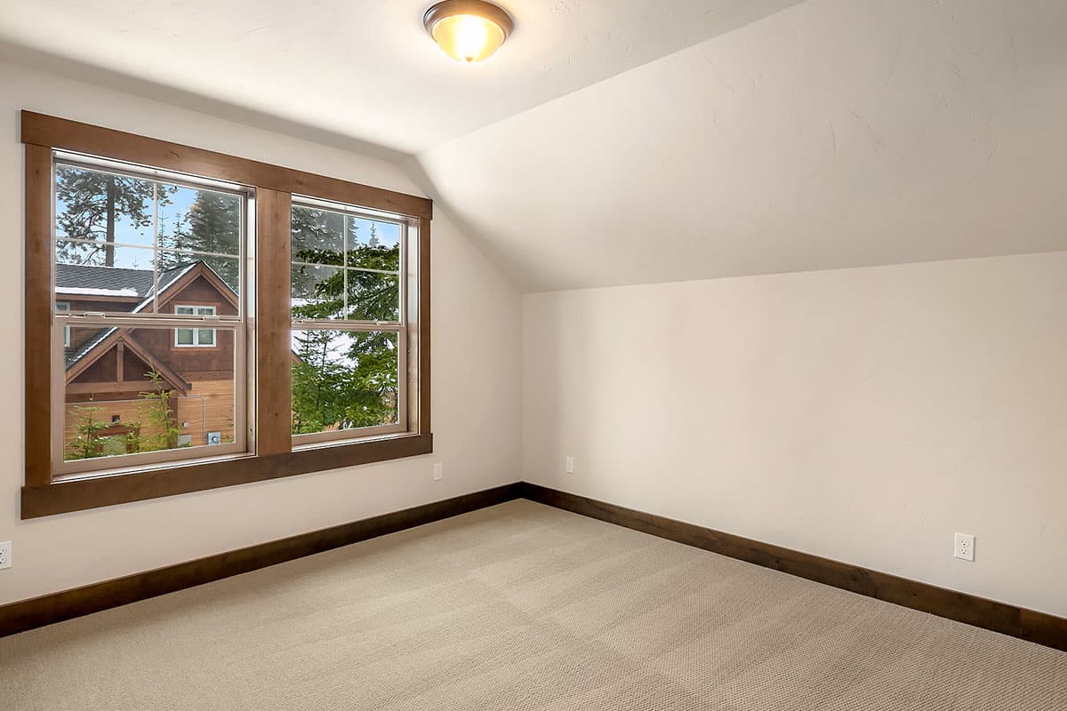 Interior view of a room with sloped ceiling, beige carpet, and two large double-hung windows with wood trim overlooking a wooden house.