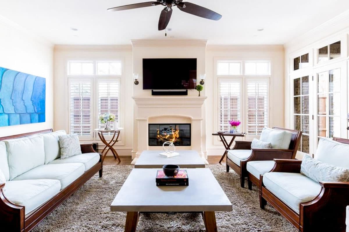 Living room interior with fireplace, television, sofas, chairs, and large windows with shutters.
