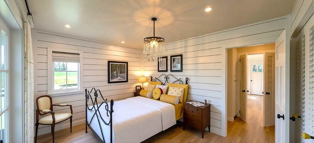 Bedroom interior with horizontal paneling on walls. Features iron bed frame, chandelier, and a view into a hallway with shuttered doors.