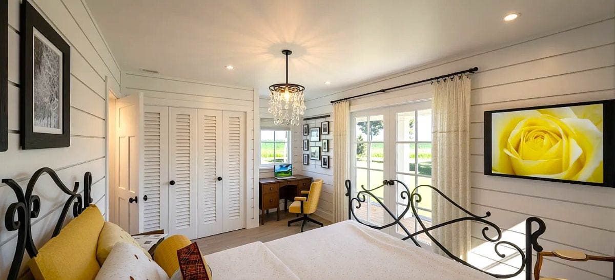 Bedroom interior with paneled walls, a chandelier, and large windows. Includes a bed, closet with louvered doors, and a small desk.