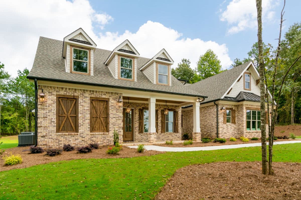 House plan exterior. Two-story brick home with gabled roof, dormers, and front porch with columns. Features wooden shutters and accent lighting.