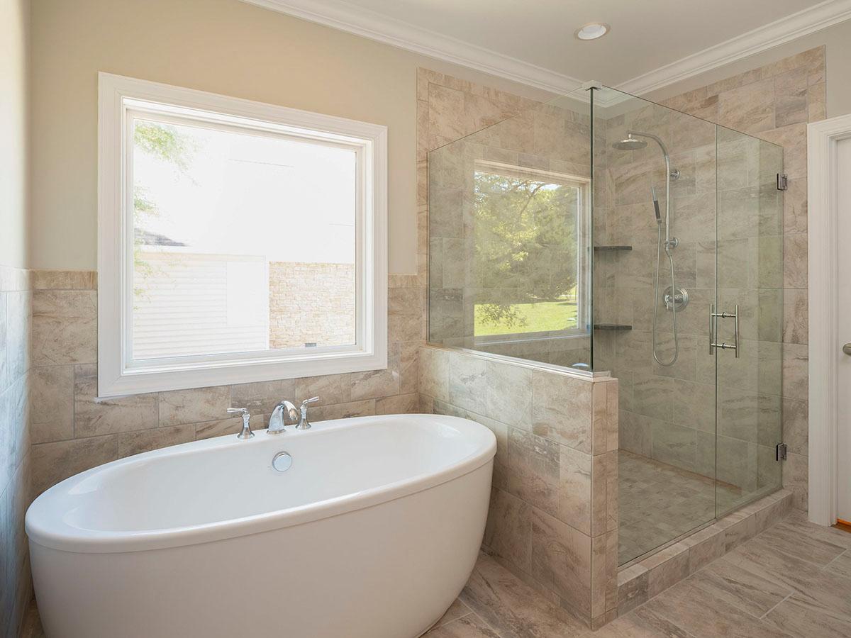 Freestanding tub and glass-enclosed shower with dual showerheads in a tiled bathroom with a large window.