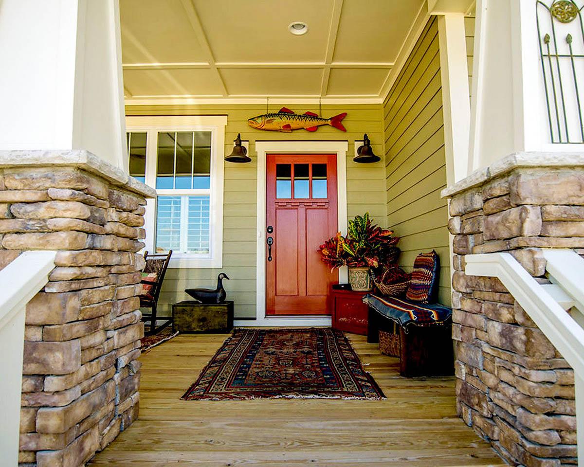 House plan exterior: Covered porch with stone pillars, wood siding, central red door, and a window.