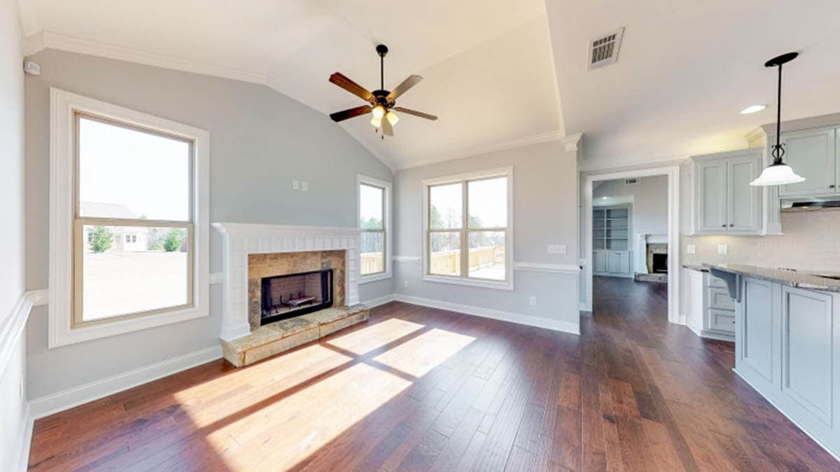 Interior view of a living room with vaulted ceiling, fireplace, and adjoining kitchen with cabinets.