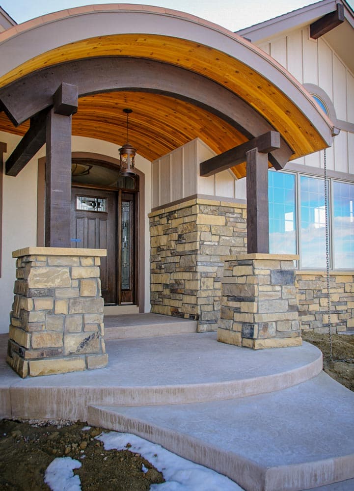 House plan exterior: Curved wooden porch with stone-clad pillars, dark wood beams, and a wood-paneled ceiling.