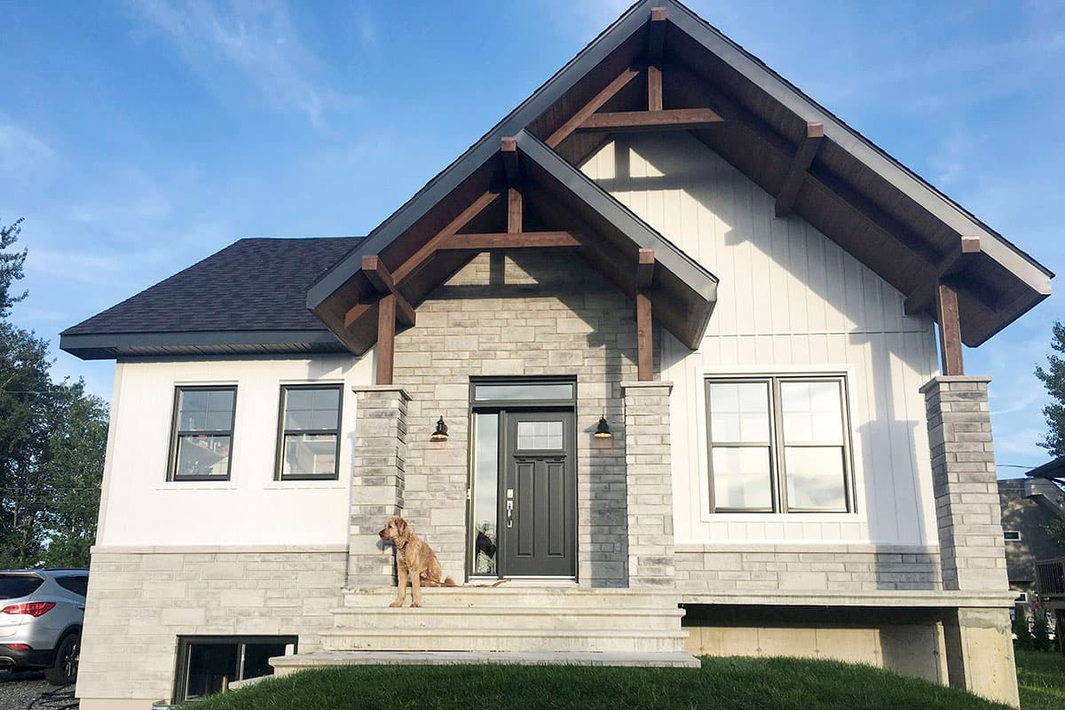 Modern house exterior with stone accents, board and batten siding, prominent timber roof supports, and a covered entry with a dark door.