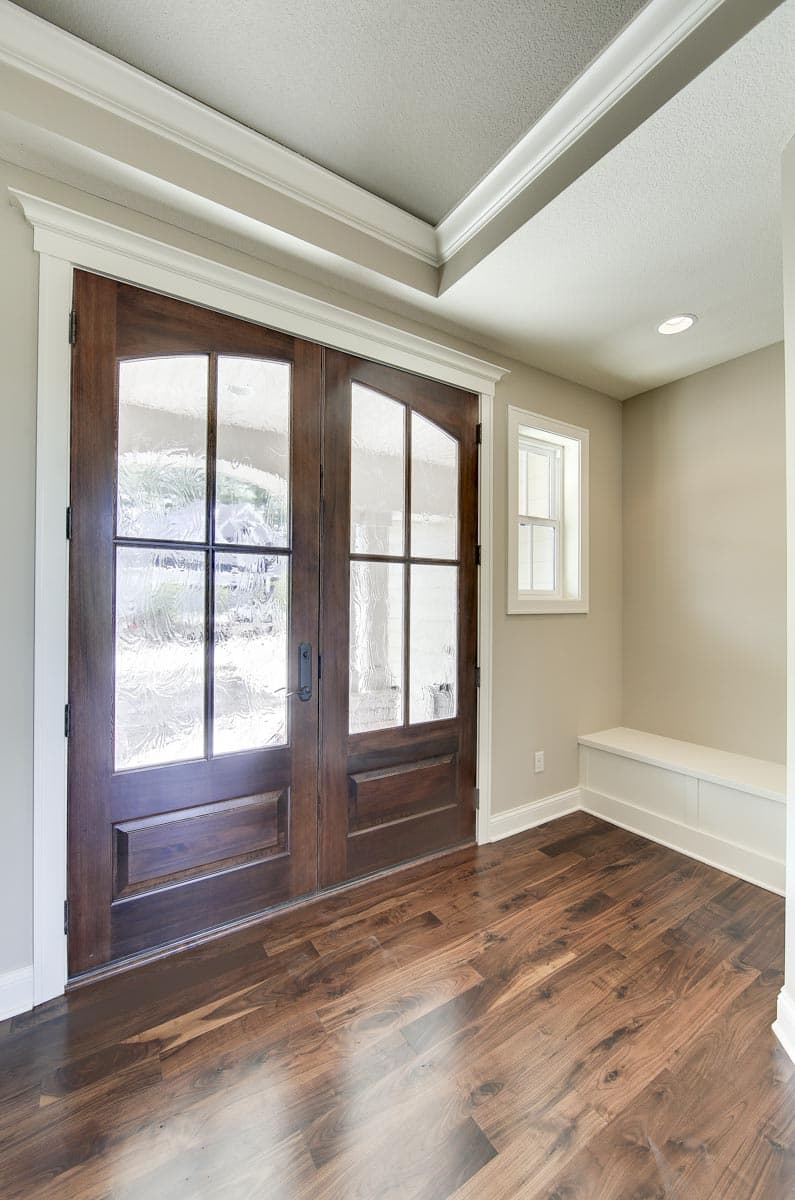 Interior view of a foyer featuring double wooden doors with glass panels, a built-in bench, and hardwood flooring.