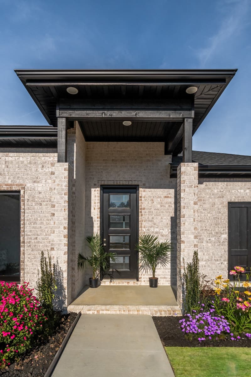 House plan exterior with light brick, modern front door with glass panes, and dark wood porch overhang.