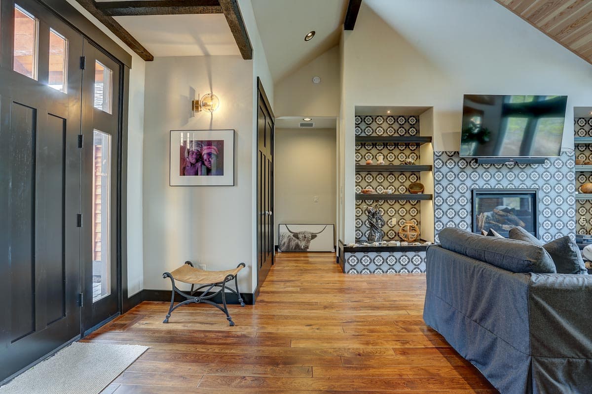 Interior view of a living room with wood floors, vaulted ceiling with beams, and a fireplace with decorative tile surround and built-in shelving.