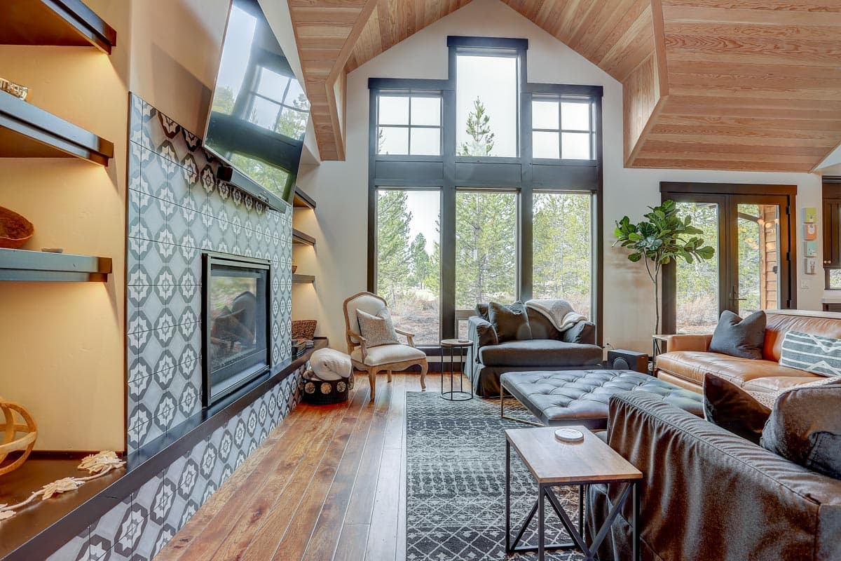 Great room with vaulted wood ceiling, fireplace, patterned tile surround, and large windows.