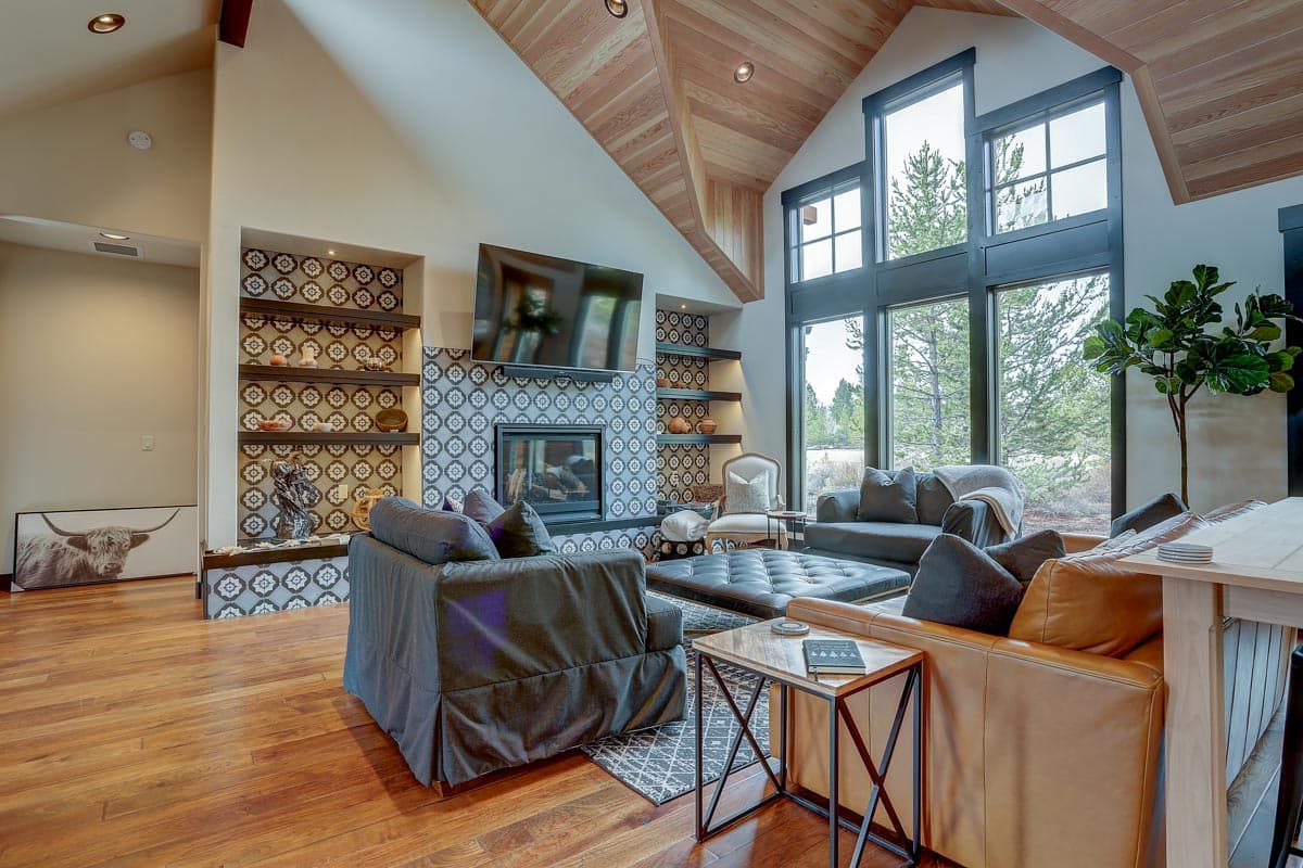 Great room with vaulted wood ceiling, patterned tile fireplace and hearth, and large windows with forest views.