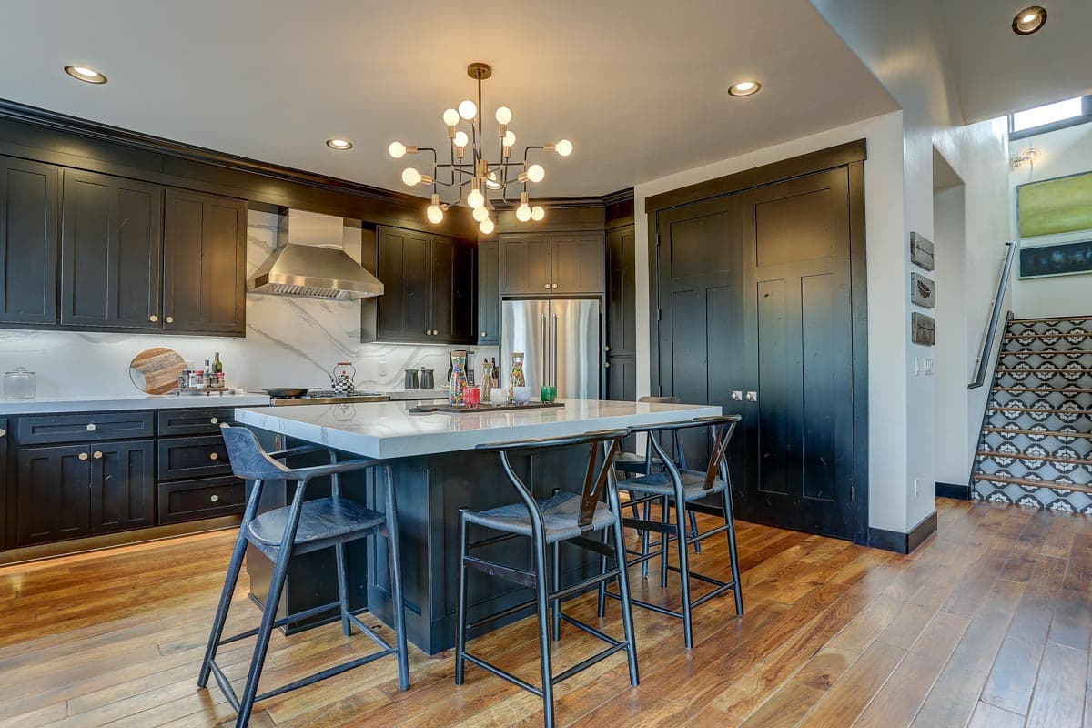 Kitchen with black cabinets, large island with marble countertop, stainless steel appliances, and decorative staircase.