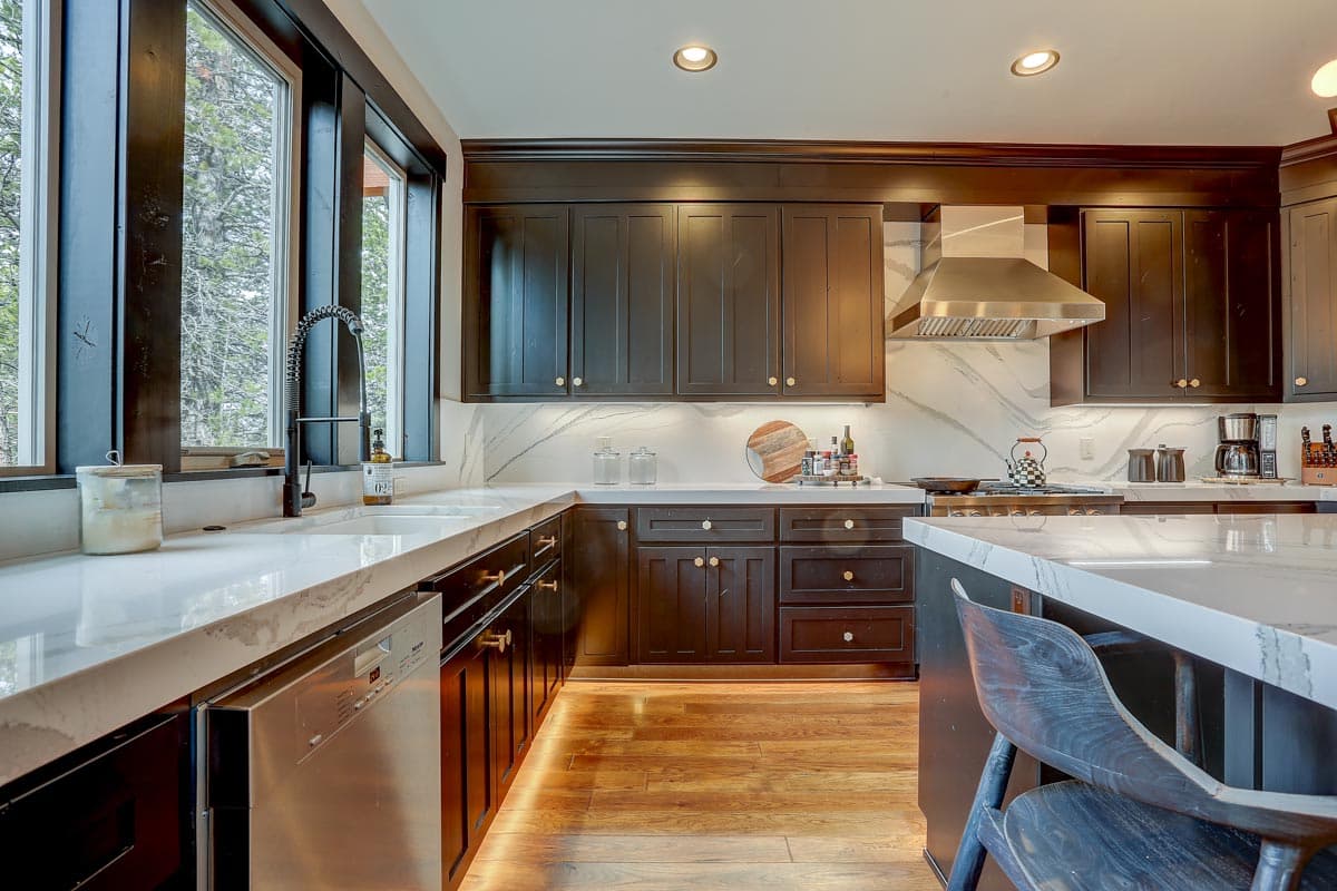 Modern kitchen with dark cabinets, white marble countertops, stainless steel range hood, and large windows.