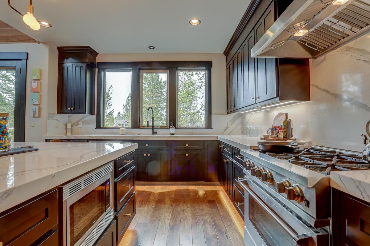 Kitchen with dark wood cabinets, marble countertops, stainless steel appliances including a range and microwave, and three large windows.