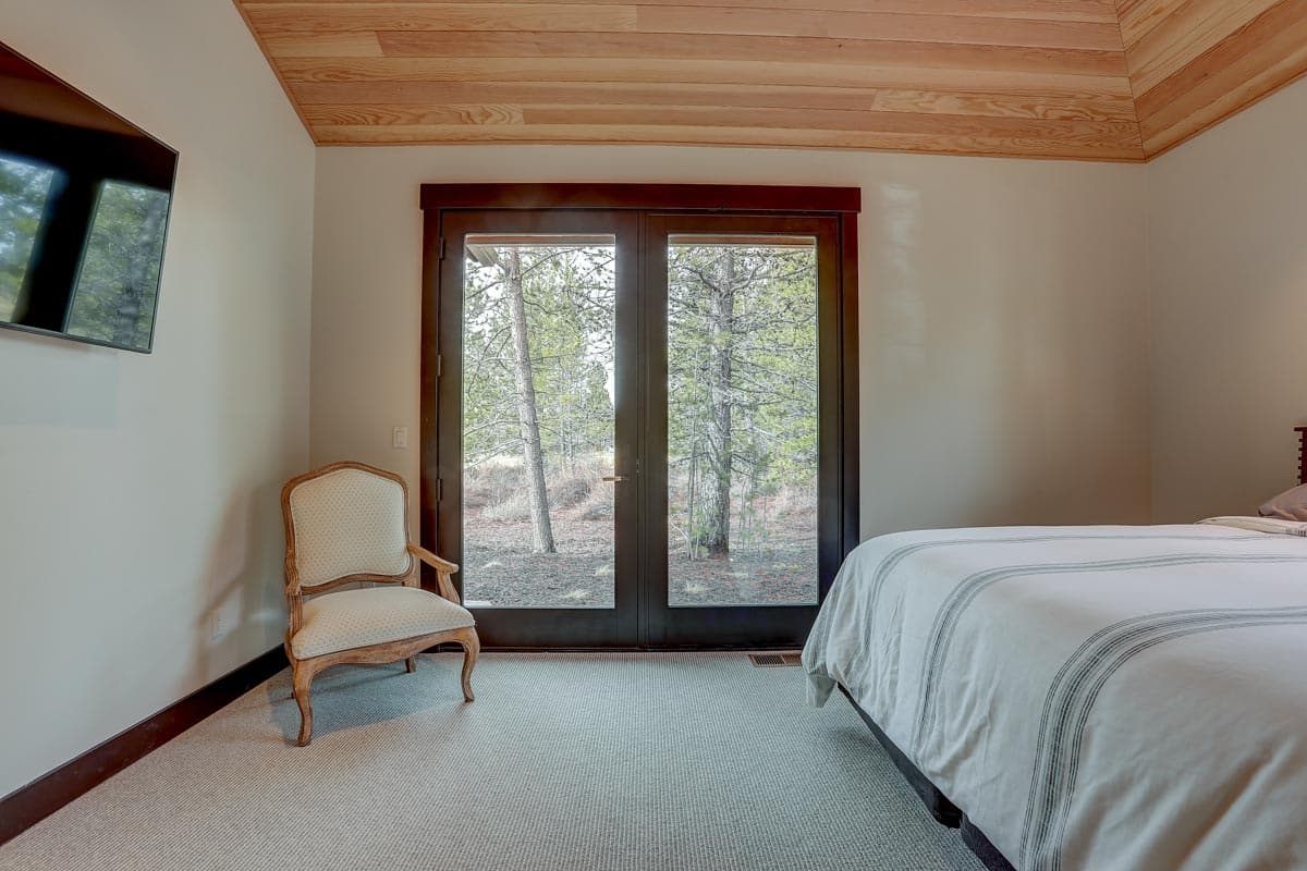 Bedroom interior with double doors opening to a wooded view, wood plank ceiling, upholstered chair, and bed.