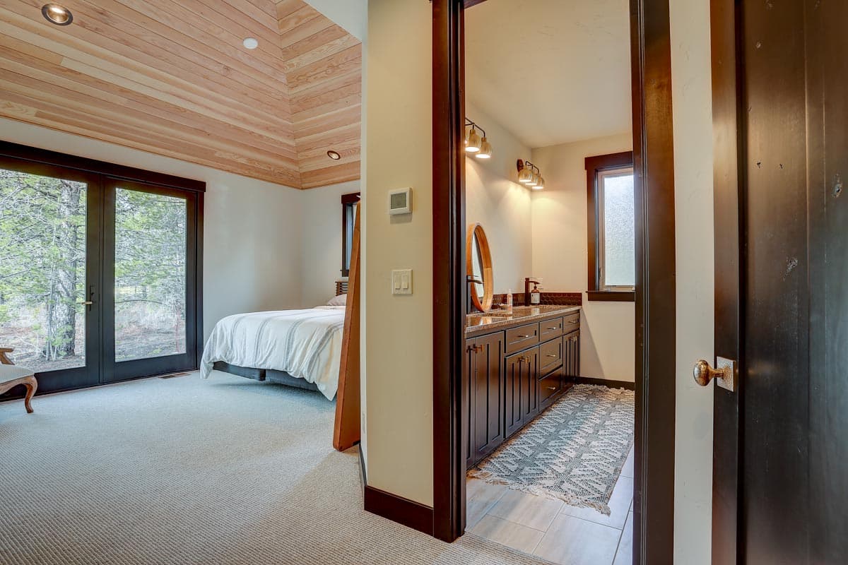 Bedroom with wood-paneled coffered ceiling and glass doors leading outside. Adjacent bathroom with double vanity and mirror.