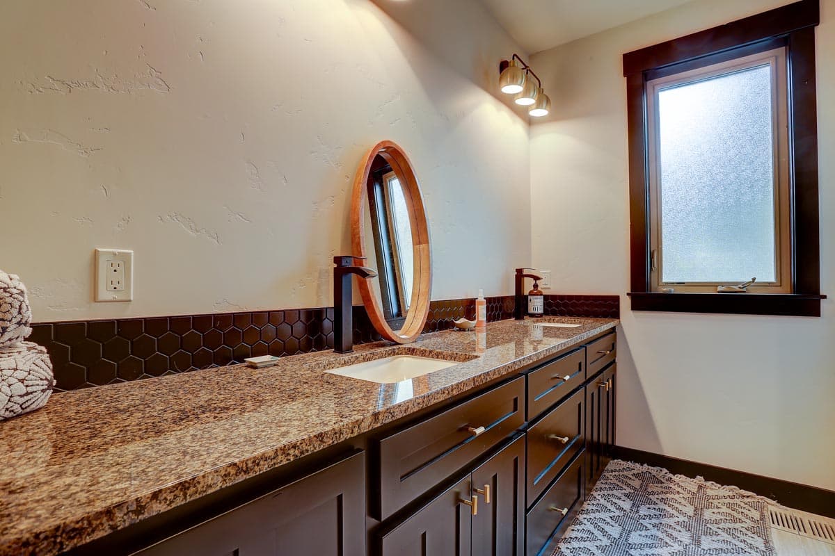 Bathroom vanity with dual sinks, granite countertop, dark cabinets, hexagonal backsplash, oval mirror, and frosted window.