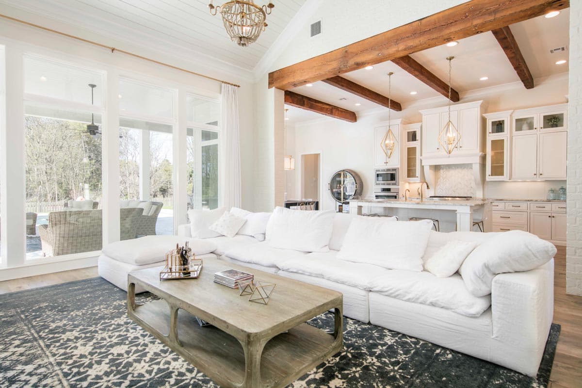 Great room with white sectional sofa, coffered ceiling with wood beams, large windows, and adjacent kitchen with island.