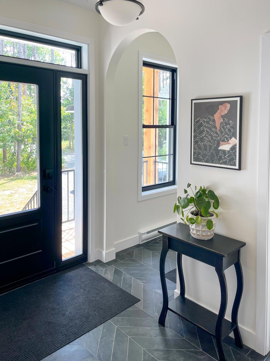 A modern entryway with a black front door and black framed windows. A small table holds a potted plant, and a framed art piece hangs on the wall.