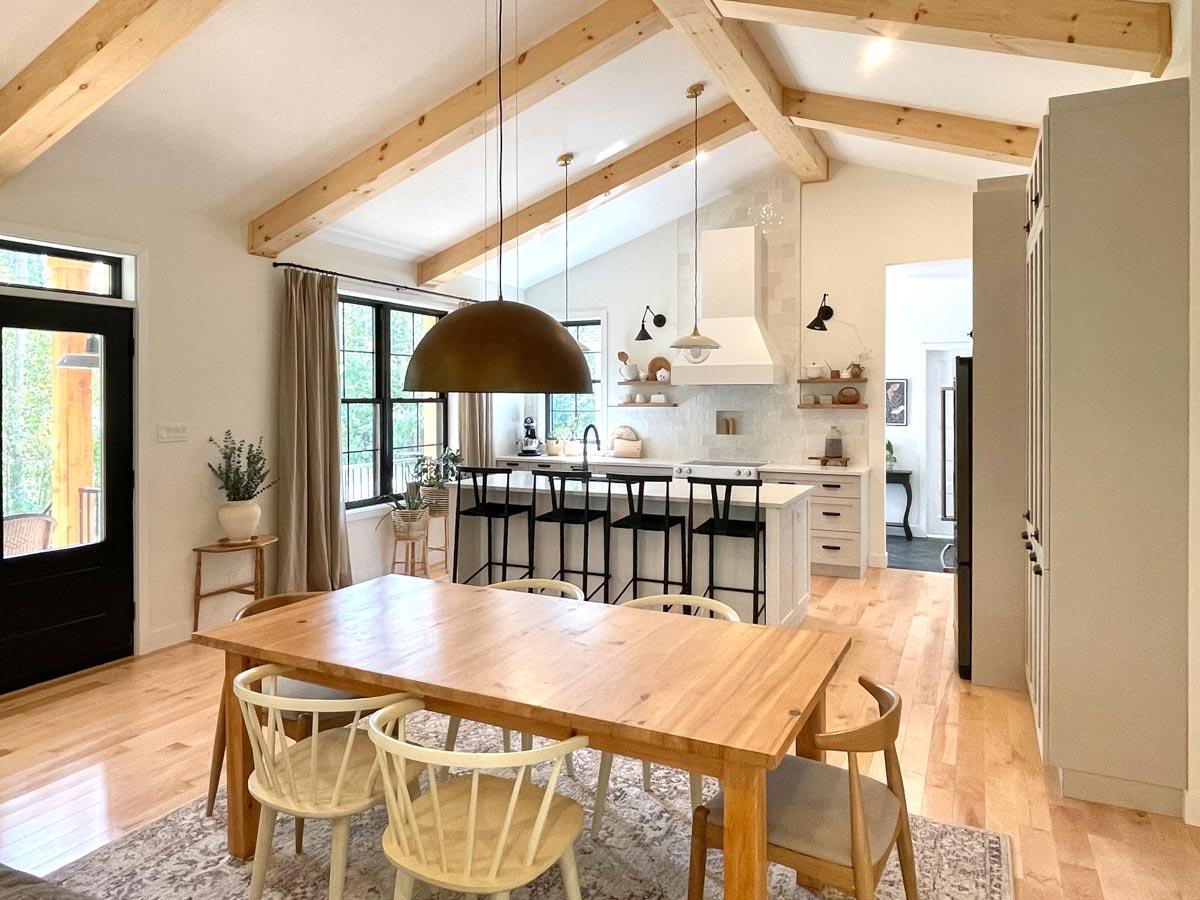 A bright, open-plan kitchen and dining area with exposed wood beams. A large wooden table with chairs sits in front of the kitchen island.