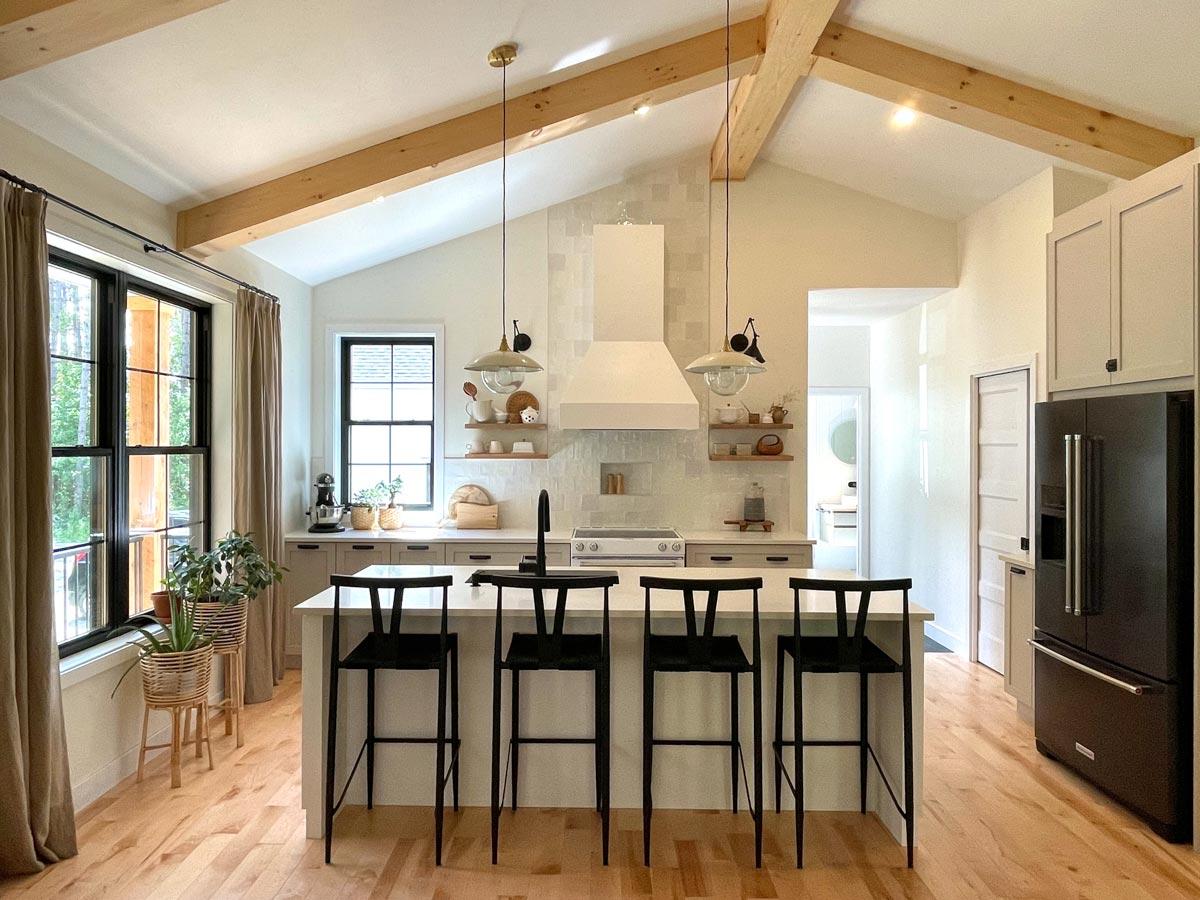 A bright, modern kitchen with hardwood floors, a breakfast bar with black stools, and a black refrigerator. Wooden beams accent the ceiling.