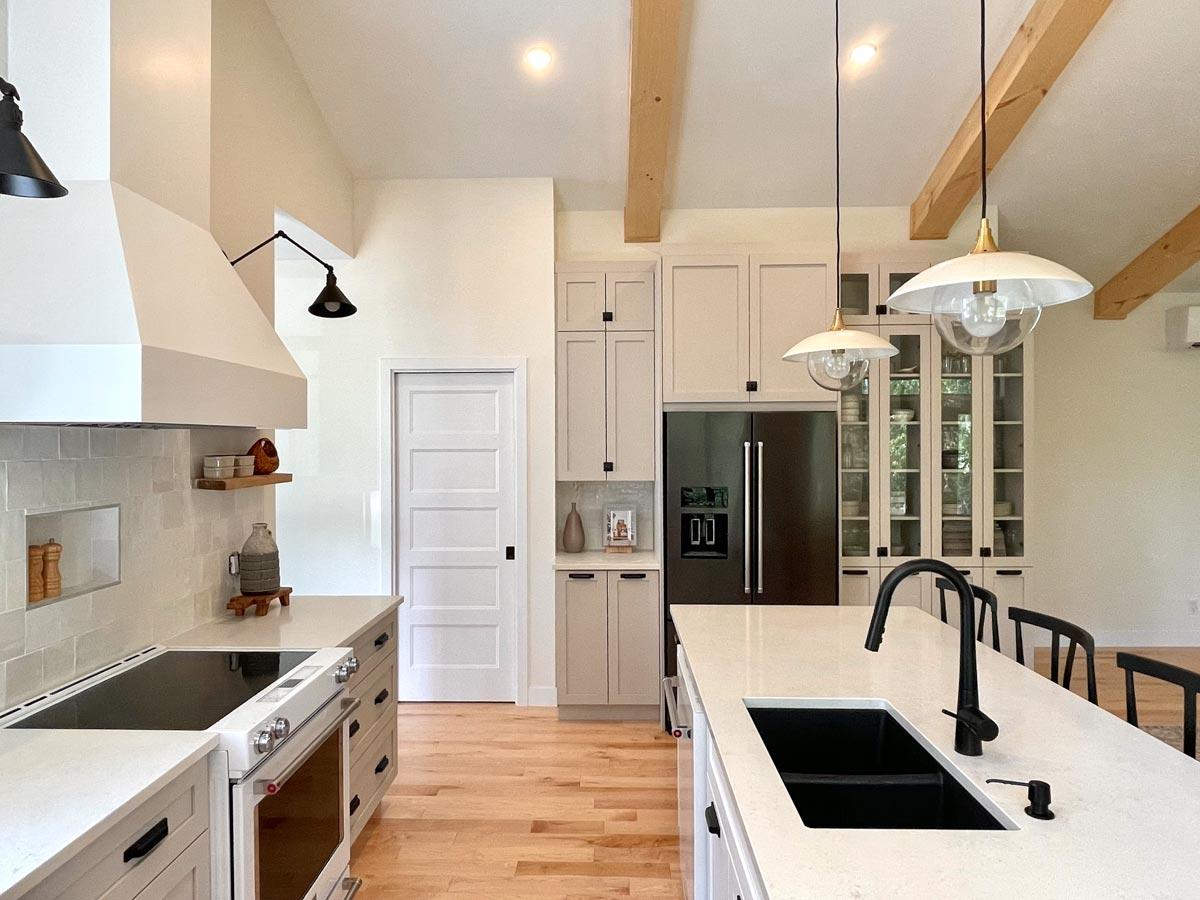 A modern, well-lit kitchen featuring a white and gray color scheme, black fixtures, and wooden floors. Overhead lights and wood beams add warmth.