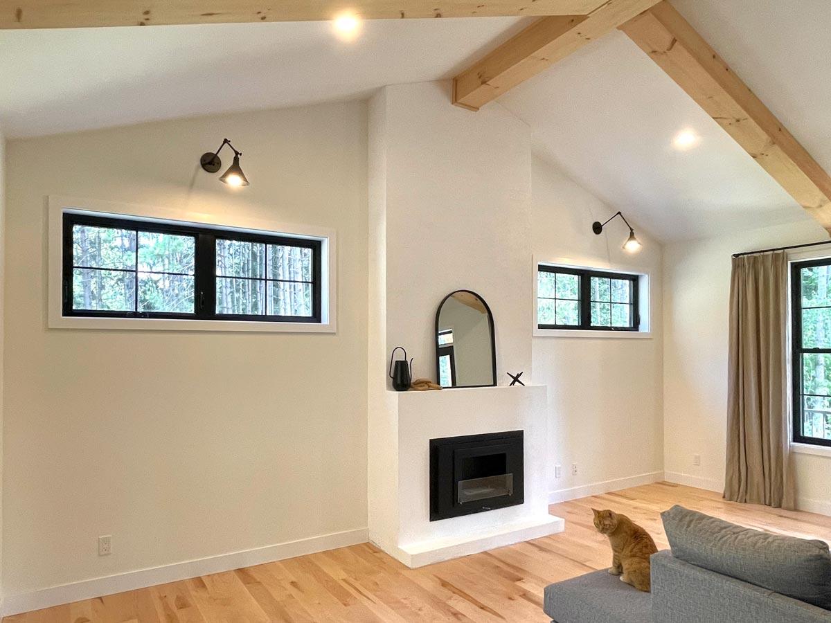 A modern, light-filled living room featuring a fireplace, black-framed windows, wood beams, and an orange tabby cat sitting on a gray sofa.