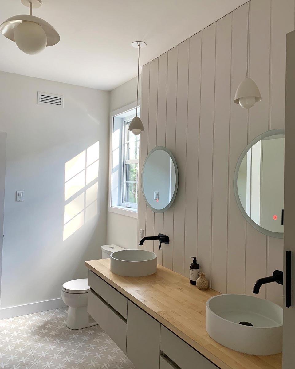 A modern bathroom features two vessel sinks on a wooden vanity with black faucets, two round mirrors, and pendant lights. Sunlight streams in from a window.