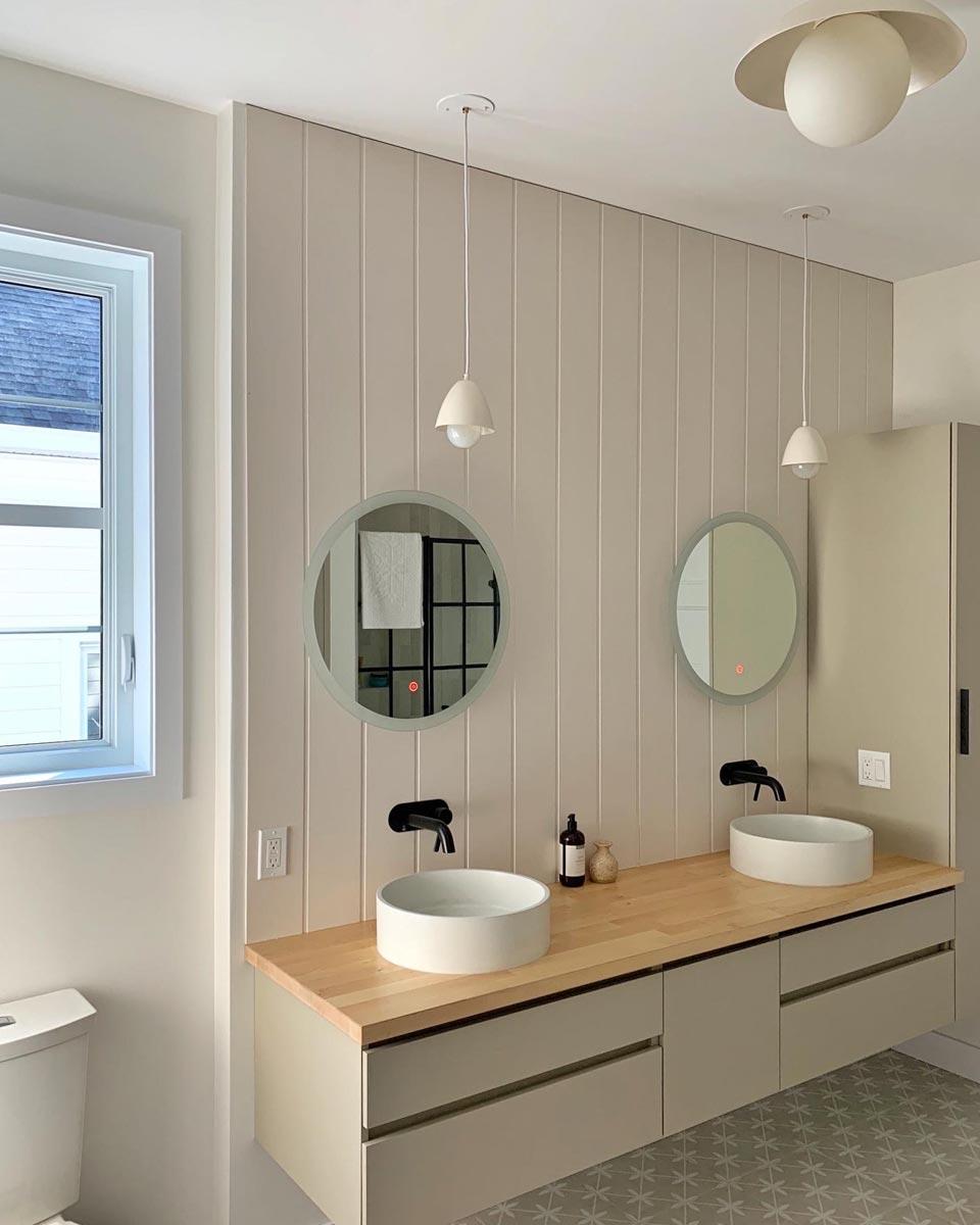 Modern bathroom featuring dual sinks, round mirrors, and pendant lights. A light-colored wood countertop sits above a gray cabinet with drawers.