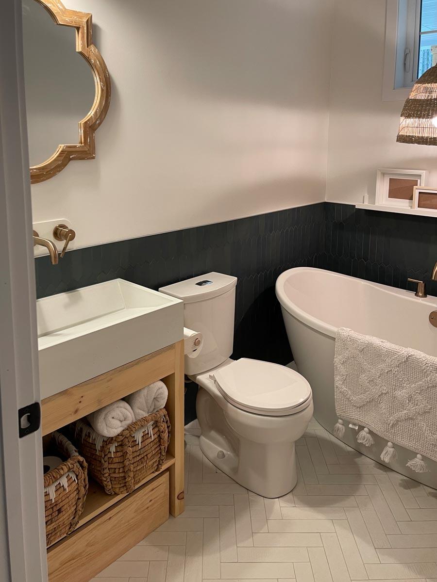 Stylish bathroom with a white tub, wooden vanity, and woven baskets. The walls are white and dark blue. A gold-framed mirror hangs above the sink.